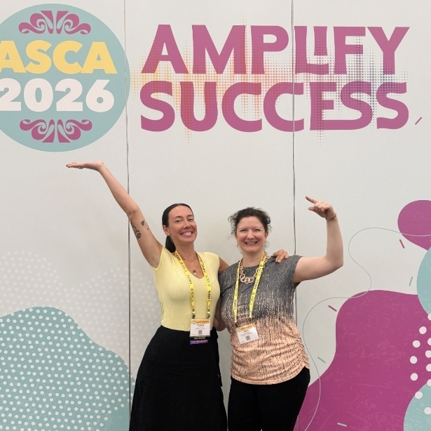 Two smiling women strike a fun pose in front of a white event backdrop featuring the "ASCA 2026" logo and the bold purple text "AMPLIFY SUCCESS." The woman on the left, wearing a yellow shirt and long black skirt, holds her right arm out and up in a playful gesture. The woman on the right, wearing a textured grey and peach t-shirt with black pants, raises her left arm and points playfully at her colleague. Both are wearing yellow conference lanyards.