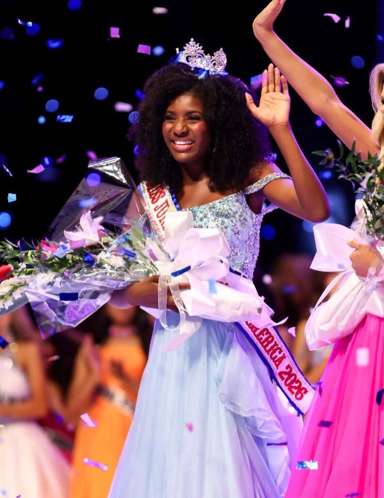 Jaidyn McDuffie wears a crown and smiles while wearing a blue dress on a stage. Confetti falls as she waves.
