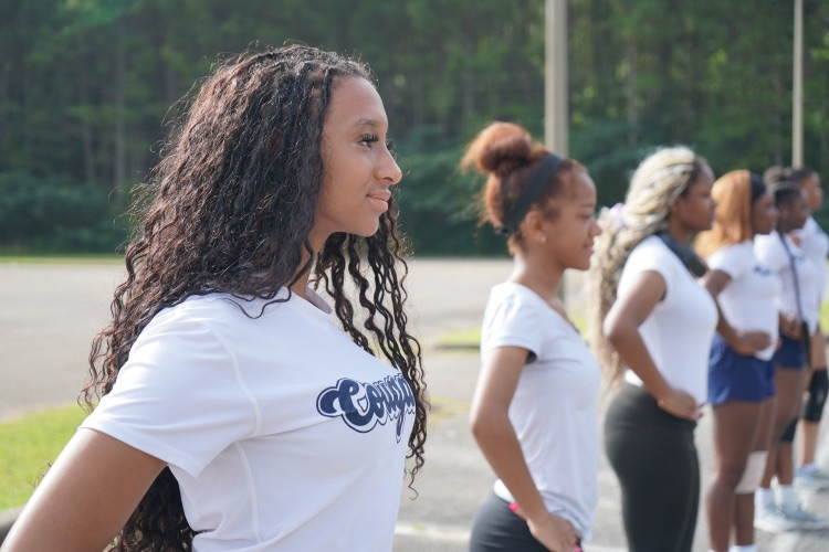 A group of girls stand in a parking lot and smile while practicing marching band fundamentals