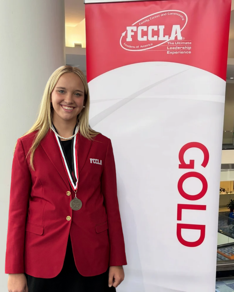 shows a student wearing red jacket and a gold medal standing next to a sign that says FCCLA Gold. 