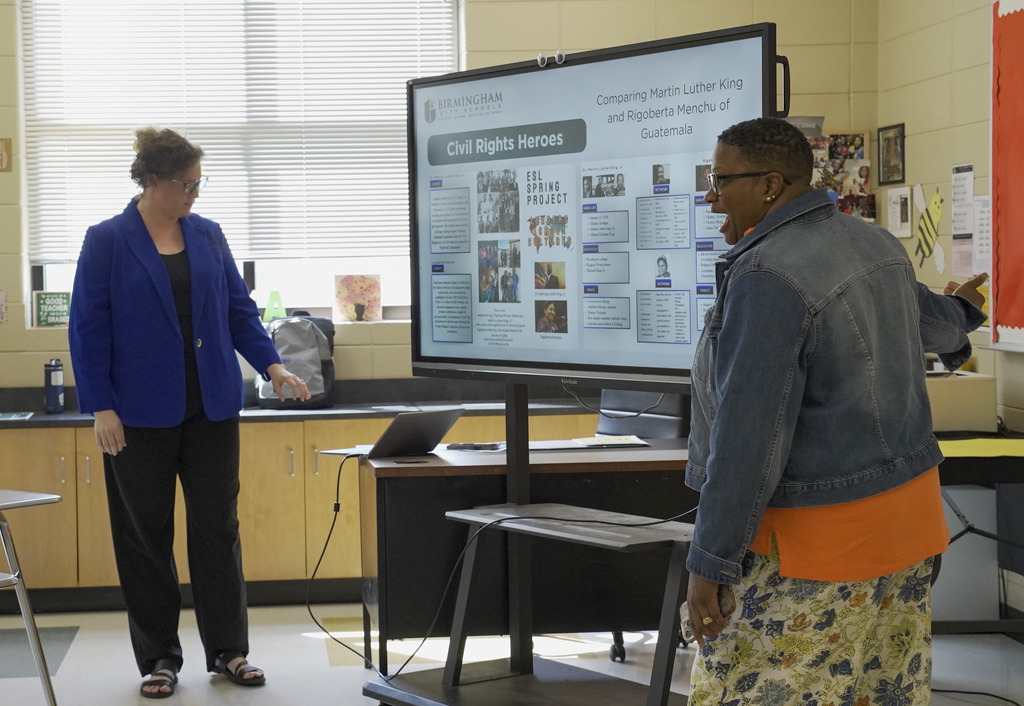 Two women stand at the front of a classroom at a smart board and lead a presentation.