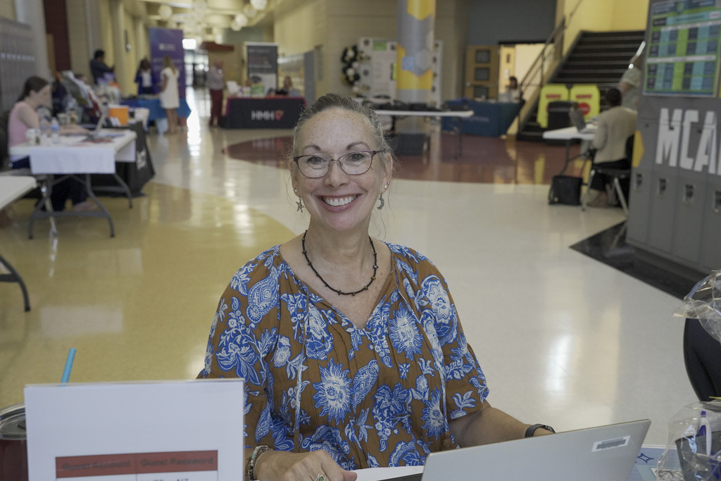 A woman sits at a table in a school hallway and smiles.