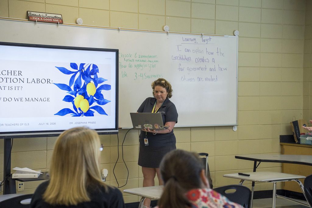 A woman stands in the front of a classroom and holds a laptop while giving a lesson.