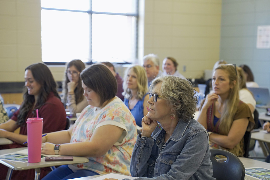 Educators sit in desks inside a classroom and listen to a speaker.
