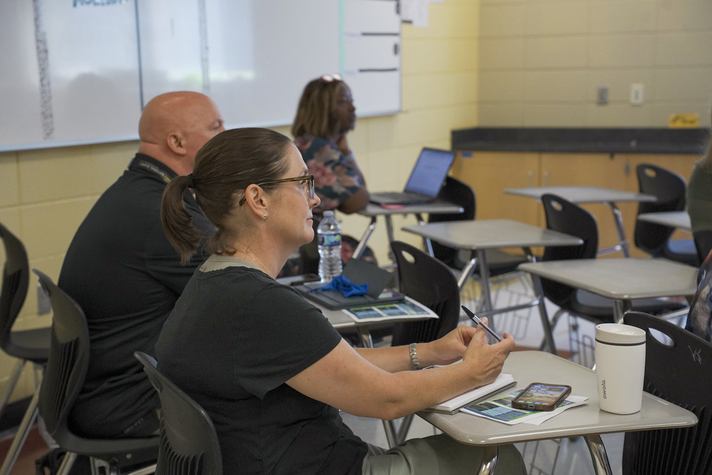 Educators sit at desks inside a classroom and listen to a presenter.