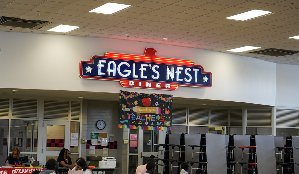 Image is of a school cafeteria. A neon sign in the cafeteria says Eagle's Nest Diner.