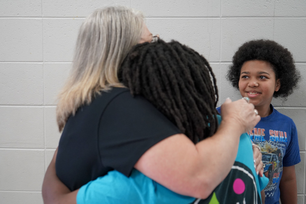 A child smiles and looks at a teacher as the teacher hugs a student. They all stand in a school hallway.