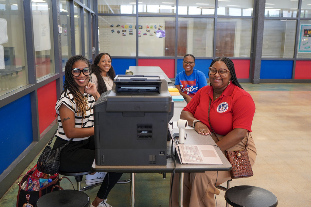 A group of women sit at a table in a school lunch room and smile.