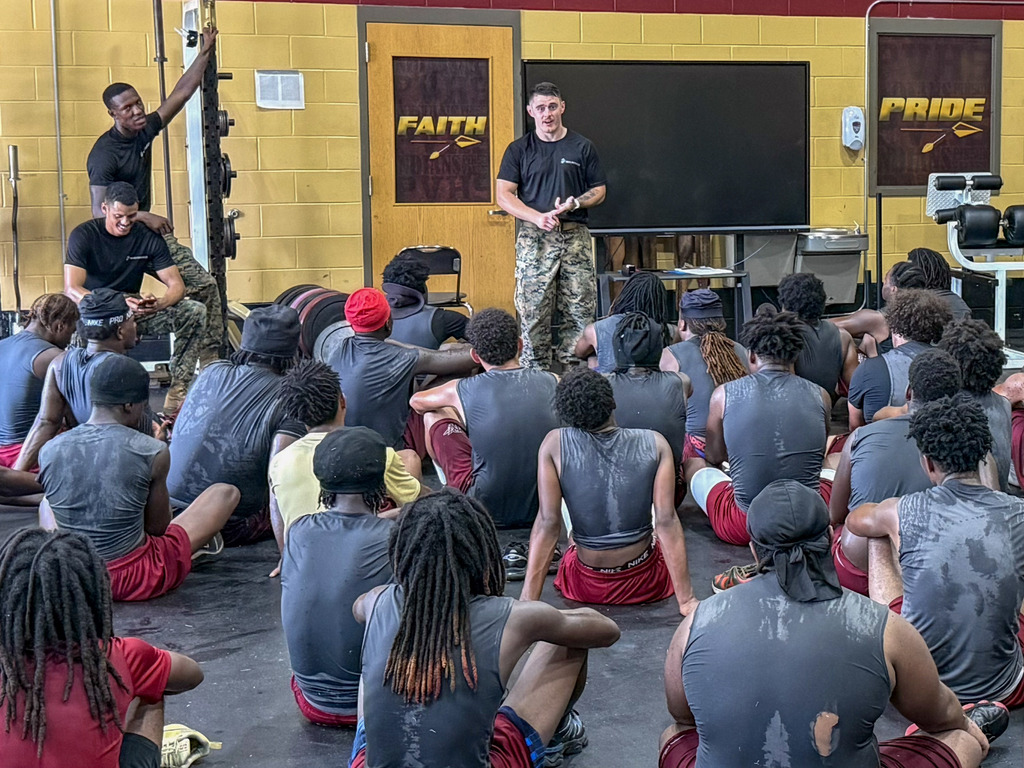 Marines stand in a football workout room and speak to Pinson Valley football players. The players sit on the floor of the room and listen.