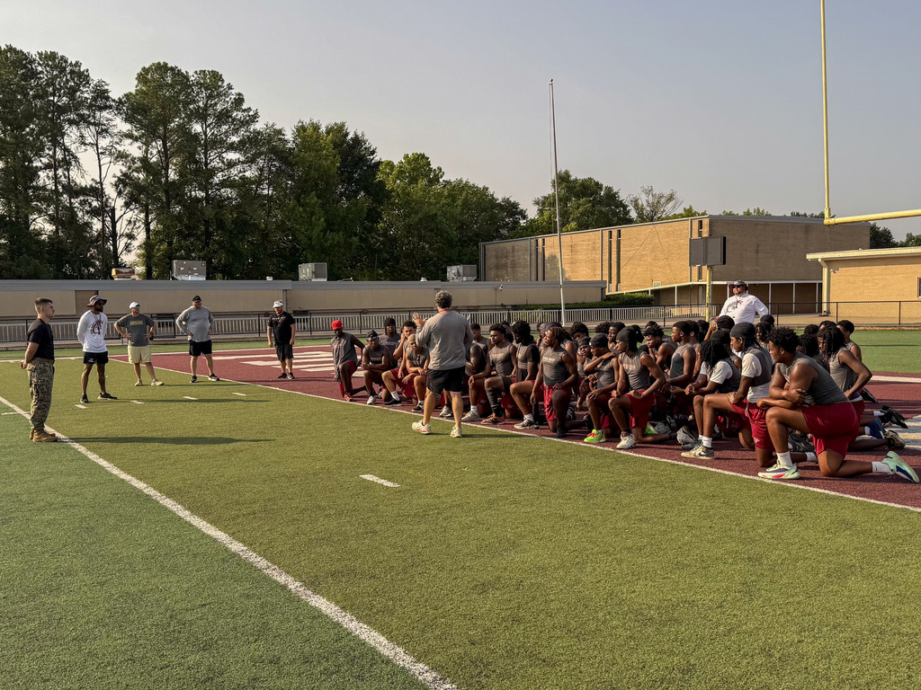 Marines stand on a football field and speak to Pinson Valley football players. The players sit on the end zone and listen.
