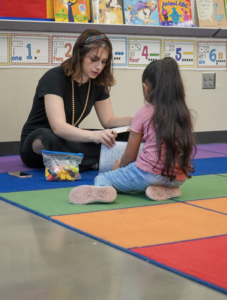 A teacher and a student sit on a colorful rug on the floor of a classroom. The teacher writes on a piece of paper while the student looks down at a worksheet.