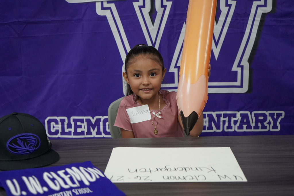 A girl smiles while sitting at a table in front of a purple backdrop and holds an inflatable pencil.