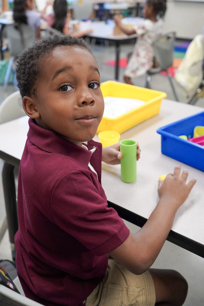 A child smiles while sitting at a table in a classroom and playing with play doh.