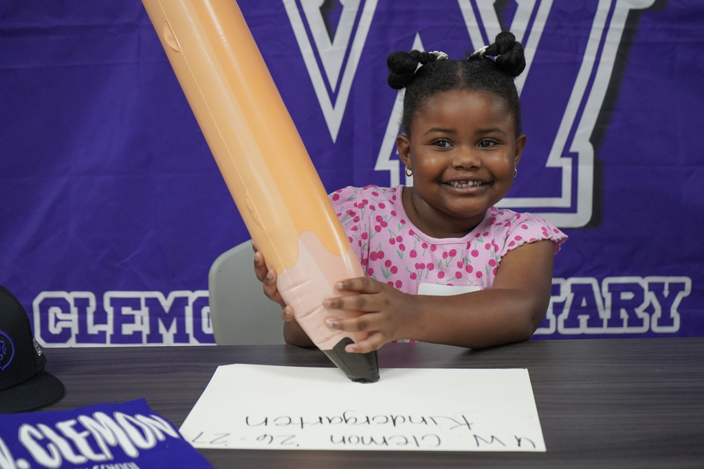 A girl smiles while sitting at a table in front of a purple photo backdrop. She holds a large inflatable pencil.
