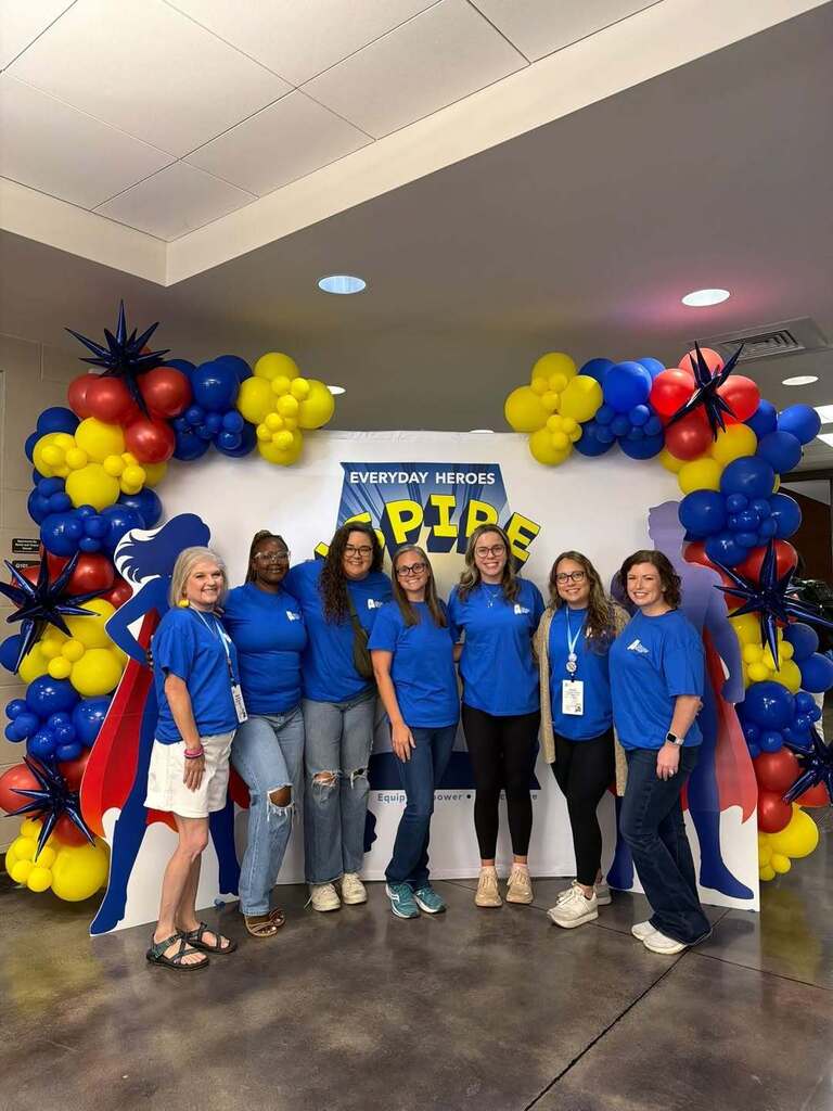 A group of women wearing matching blue shirts stand in front of a photo backdrop with balloons in a hallway and pose for a photo.