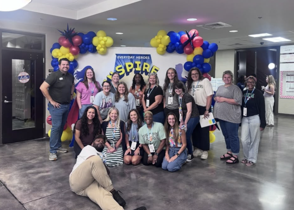 A group of teachers stand together and smile for a photo in a hallway. They pose in front of a photo backdrop with balloons on it.