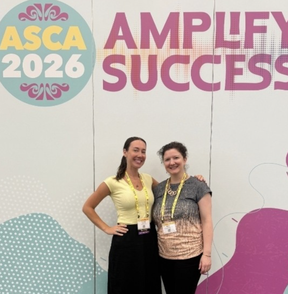 Two women pose for a photo in front of a sign. The sign says "ASCA 2026 Amplify Success."