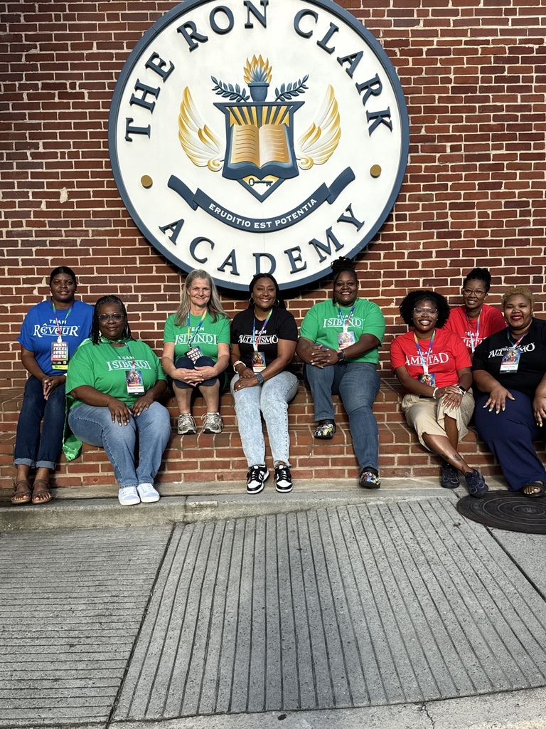 Educators sit outside a brick building. They pose for a photo under a sign on the building that says "The Ron Clark Academy."