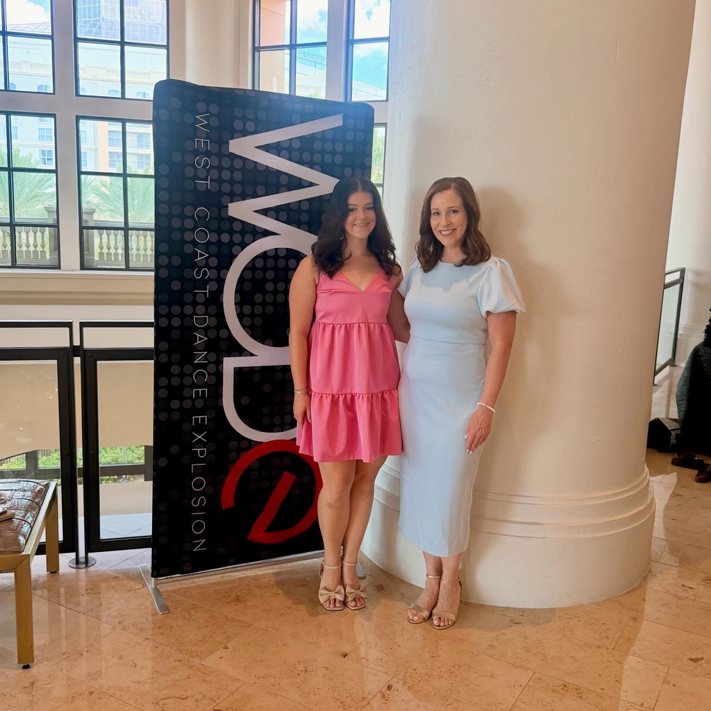 A girl and a woman stand together for a photo. They wear dresses and pose in front of a sign for West Coast Dance Explosion Convention and Competition.