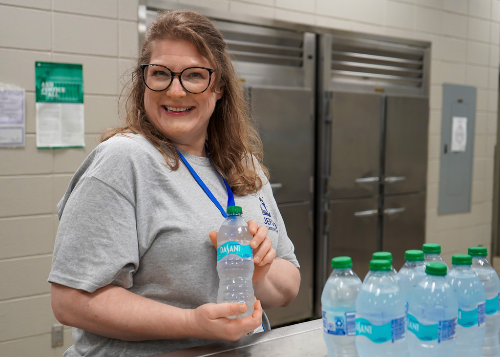 A woman stands in a kitchen and smiles at the camera while holding up a water bottle. 