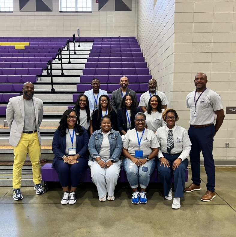 Eleven administrators representing the Minor feeder pattern in Jefferson County Schools pose for a group photo in a gymnasium at the Jefferson County Schools Administrators conference. The group features staff seated and standing in rows in front of purple and yellow bleachers, with many wearing blue lanyards and district-branded shirts.