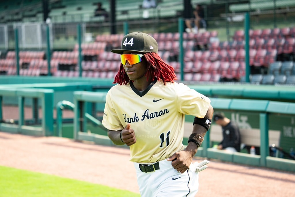 A baseball player runs across a baseball field. He wears a jersey with the number 11 and "Hank Aaron" on it. His hat has the number 44 on it.