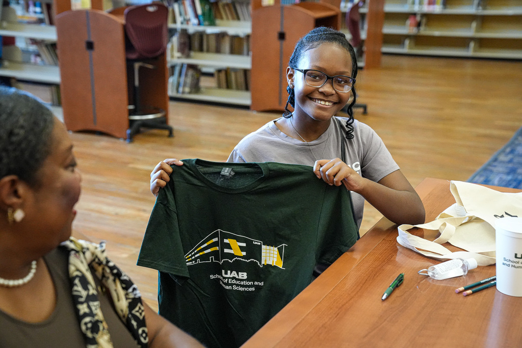 A girl sits at a table in a library and holds a UAB shirt.