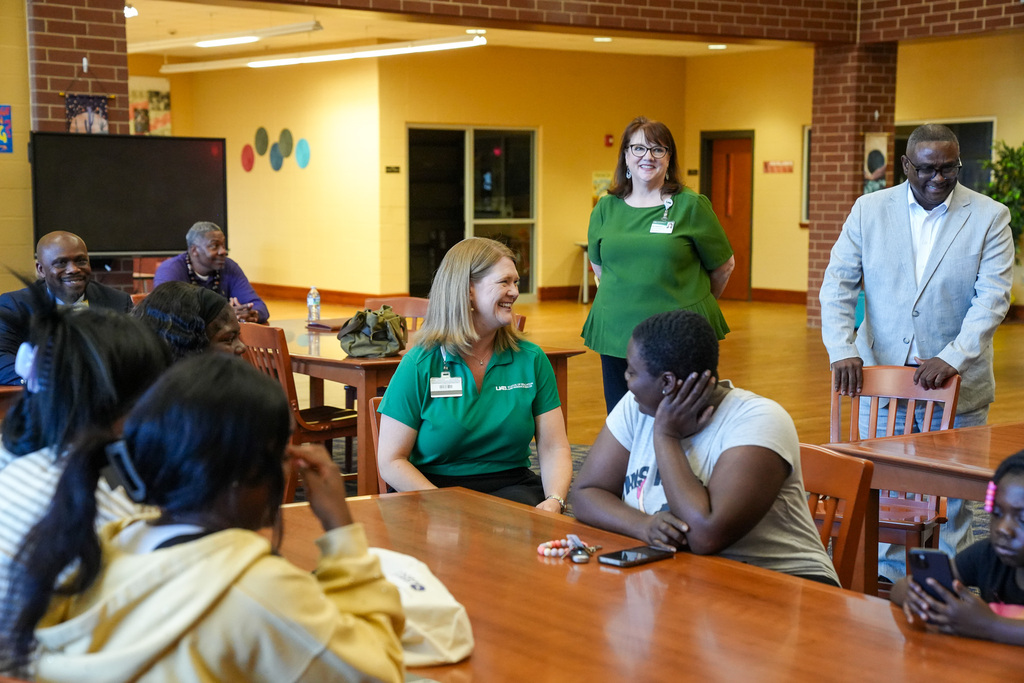 A group of adults sit at tables inside a library and speak to each other.