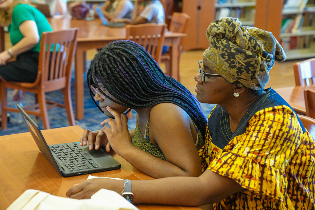 A girl and a woman sit at a table in a library. The girl looks at a laptop screen.
