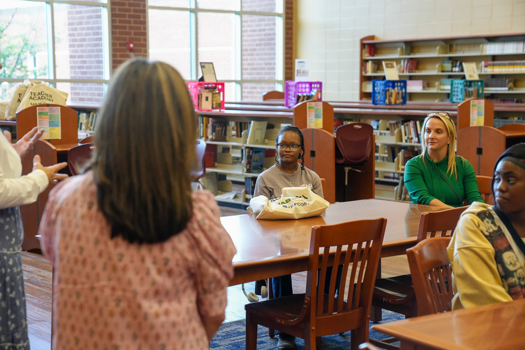 Women stand in a library and speak to a group of individuals as they sit at tables.