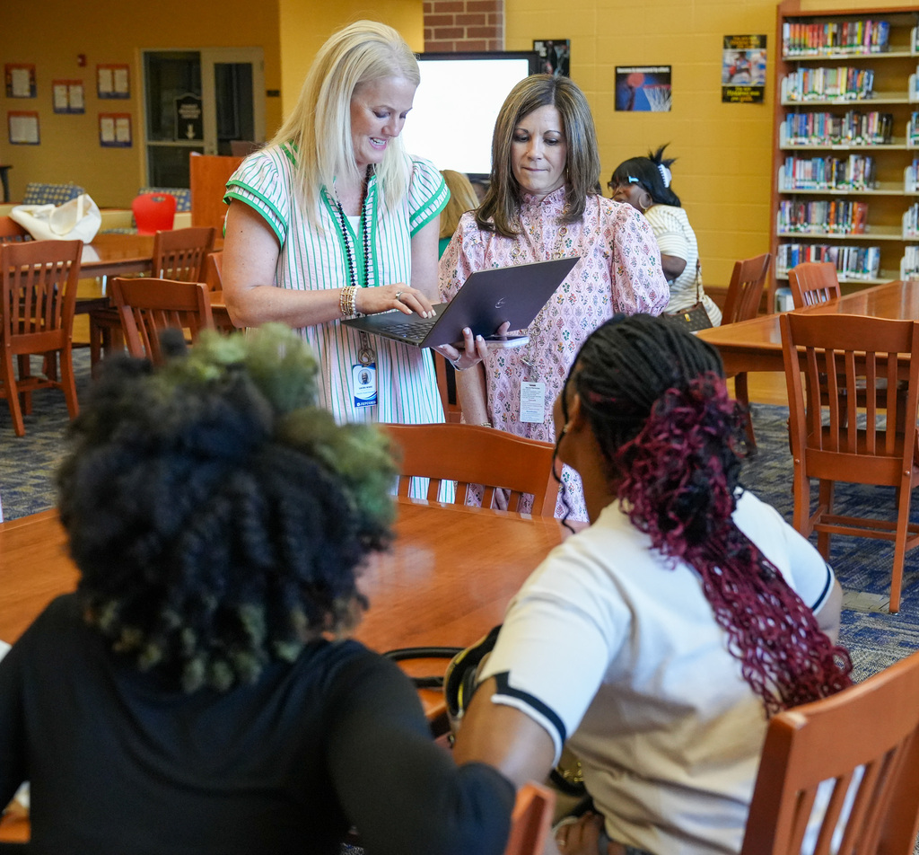 Two women look down at a laptop while standing in a library. Other people sit around them at tables.