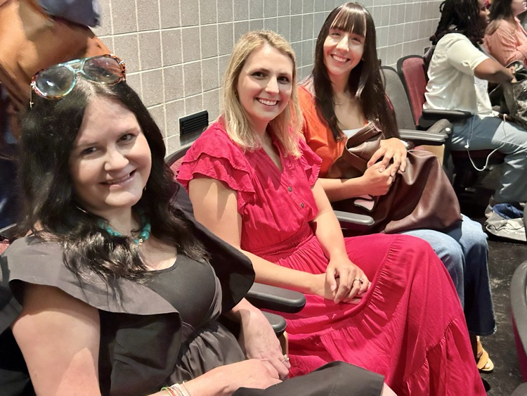 Three women sit in an auditorium and smile for a photo