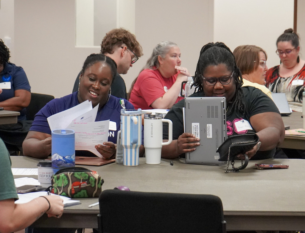 Adults sit at tables inside a room. Two women in the center of the photo smile.