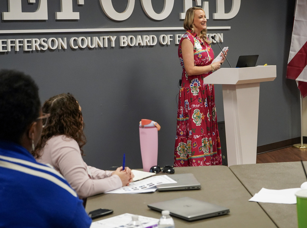 A woman smiles while standing at a podium inside a room. Educators sit at tables around her and take notes.