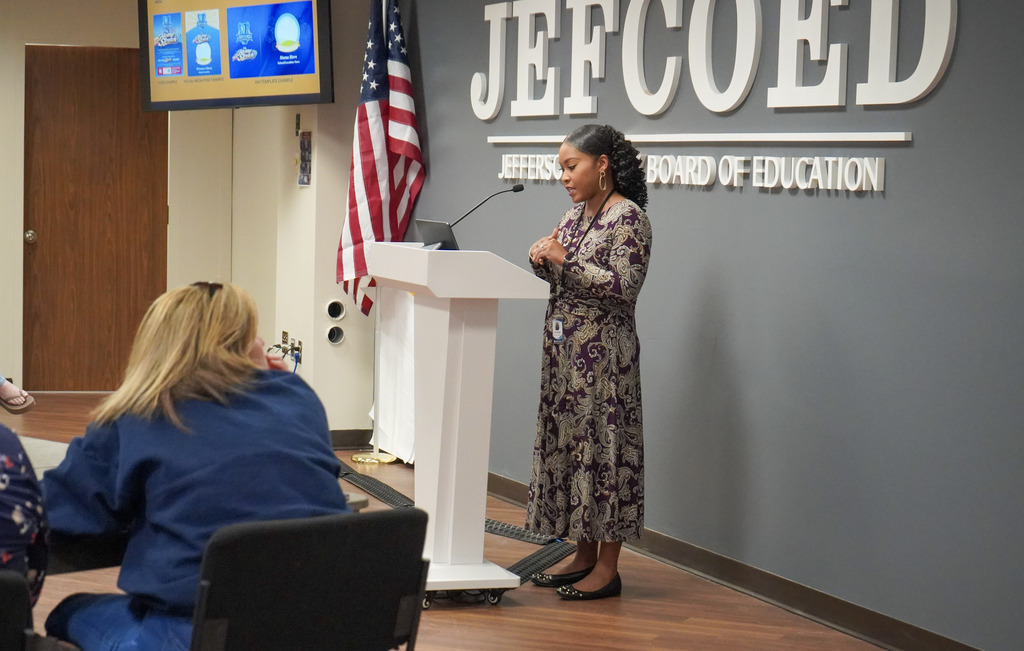 A woman stands at a podium in a room and speaks into a microphone. People sit in chairs around her and listen.