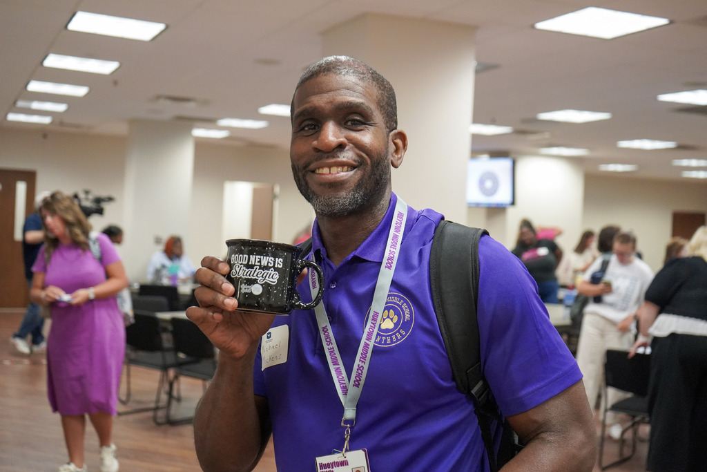A man stands in a room and smiles. He holds a coffee cup in his hand that says "Good News is Strategic."