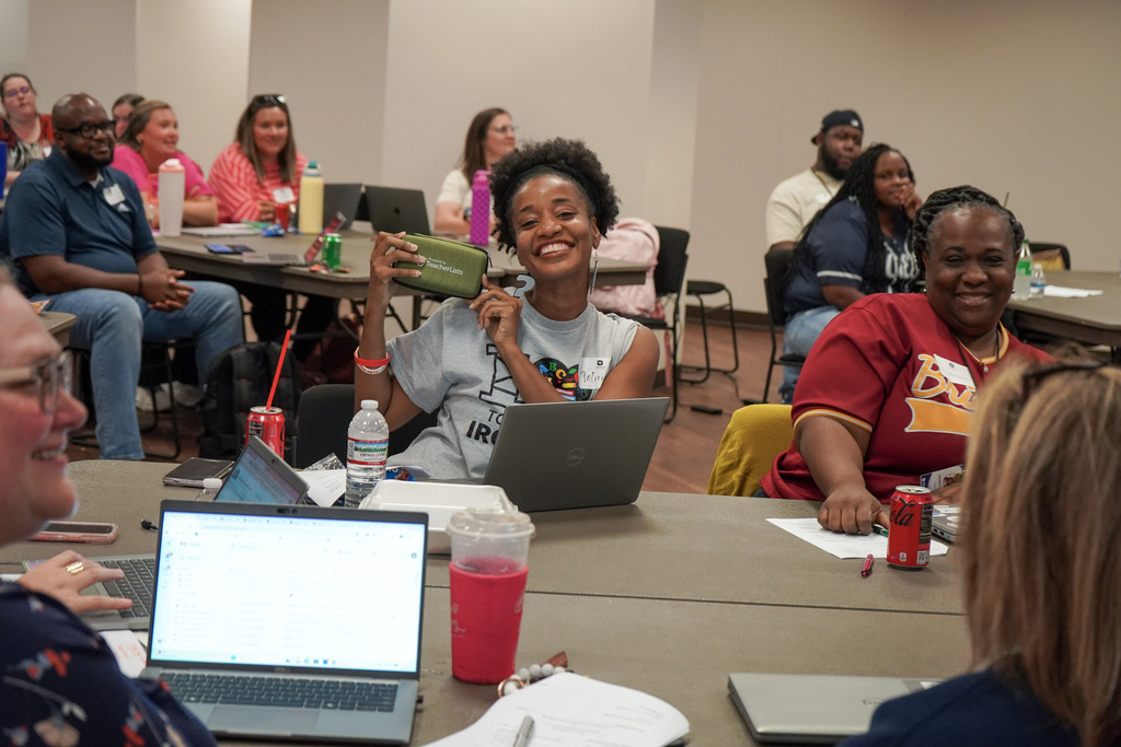 Educators sit at tables in a room and smile. One of the educators holds up a small bag.