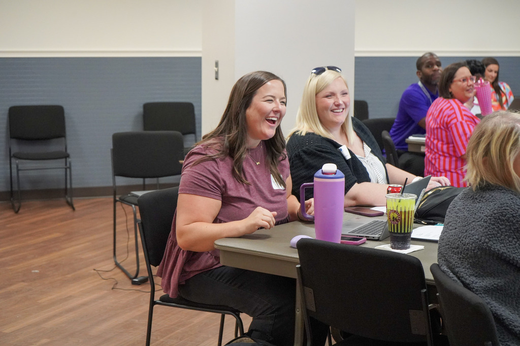 Two women smile while sitting at a table inside a room.