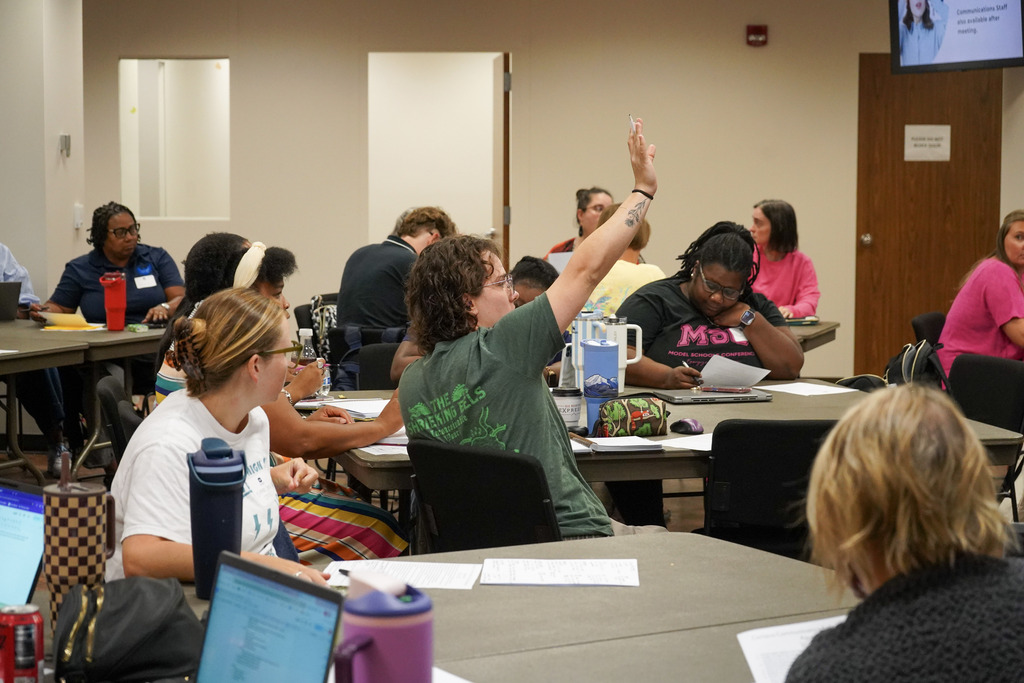 Adults sit at tables inside a room. One of the adults raises their hand.