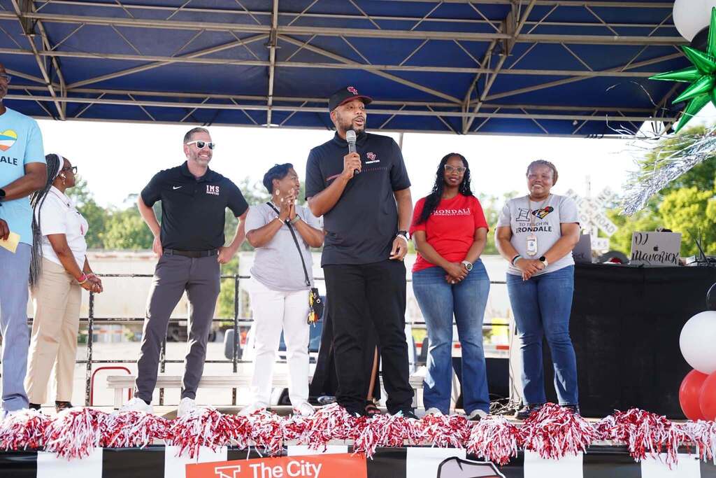 A man speaks into a microphone while adults stand behind him on a stage set up outdoors.