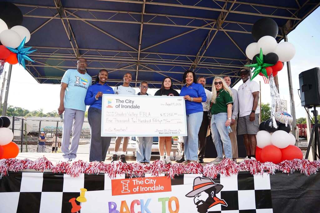 People stand on a stage outdoors and hold up a big check from the City of Irondale made out to the Shades Valley FBLA.