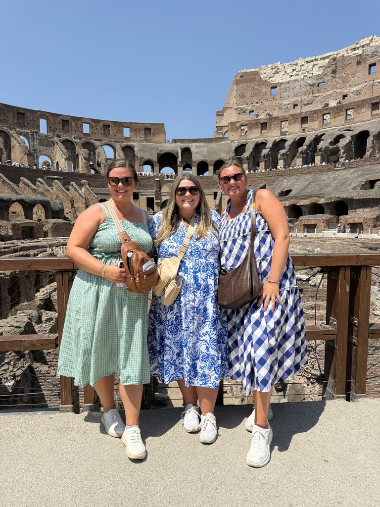 Three women smile and pose for a photo in front of the Colosseum in Rome.