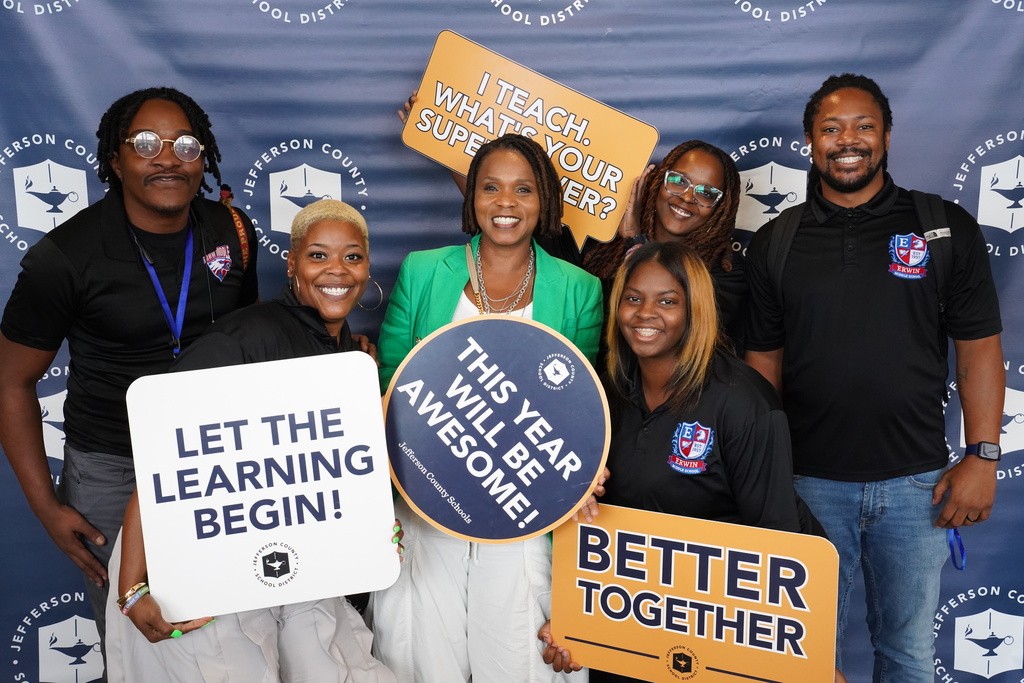 A group of adults stand together and smile for a photo. They hold education themed signs and stand in front of a Jefferson County Schools photo backdrop.