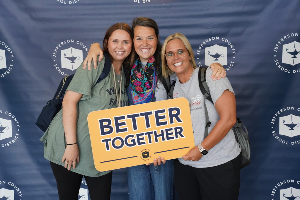 Three women stand together for a photo and smile. They stand in front of a Jefferson County Schools themed photo backdrop and hold a sign that says "Better Together."