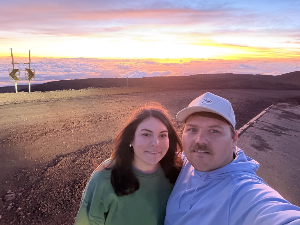 A man and a woman smiling in front of a pink, gold, and blue sky from atop a mountain.