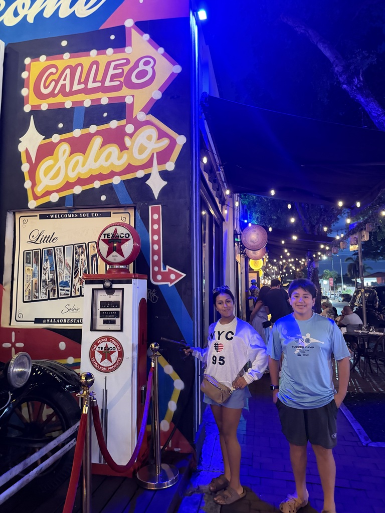 Two children stand outdoors at night. They're surrounded by bright signs and glowy lights.