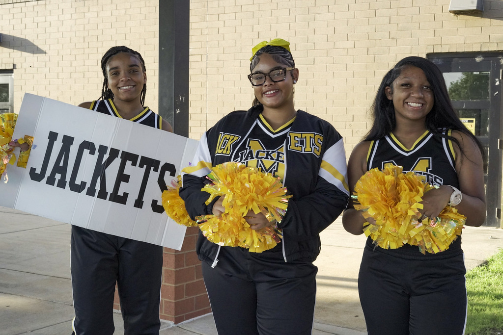 Cheerleaders hold pom poms and a Jackets sign outside a school.