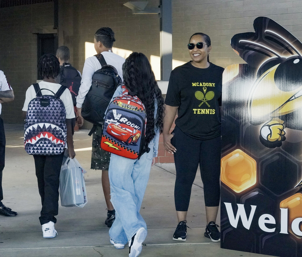 A teacher smiles while students walk past her toward the school entrance.