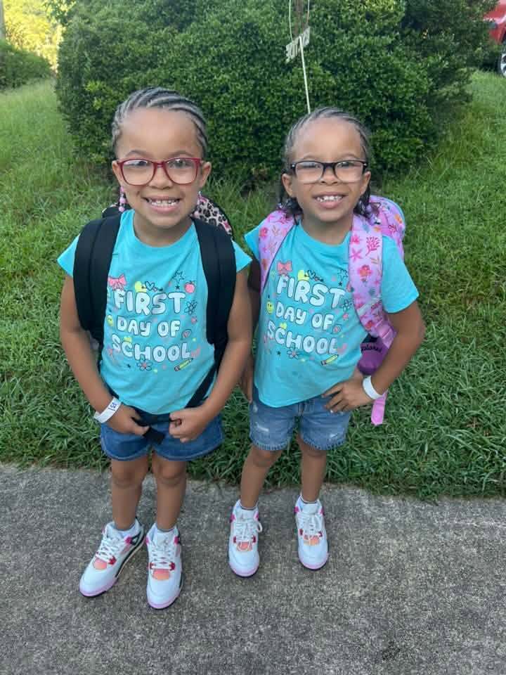 Two children wearing matching first day of school shirts stand outdoors and smile for a photo.