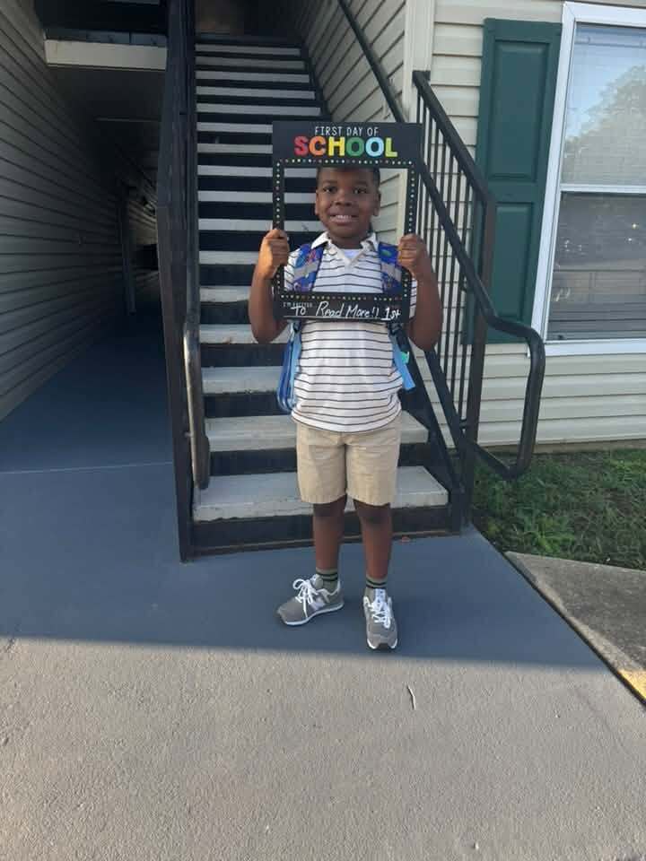 A child stands outside a building and poses for a first day of school photo.